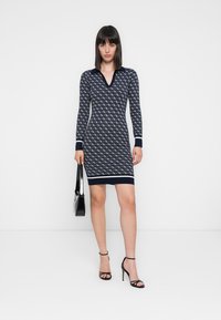Abito in maglia di colore blu navy con un motivo a diamante, scollatura a V, maniche lunghe e orlo a righe. Abbinato a una borsa nera e tacchi a strappo.