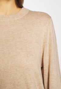 Gros plan sur une personne portant un pull beige côtelé à col rond, montrant les détails de la partie supérieure de la poitrine et des épaules.
