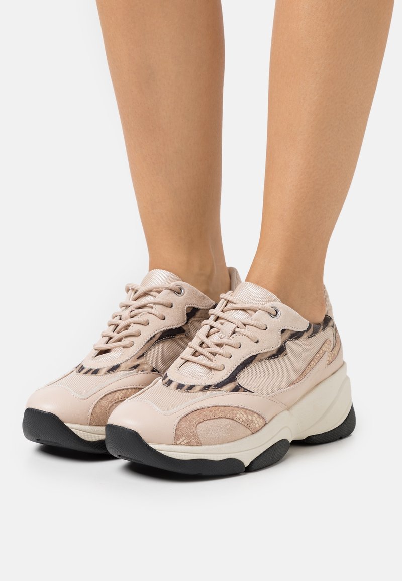 Pieds portant des baskets beige épaisses à lacets à l'avant et semelles noires, avec des détails en mesh et imprimé animal.