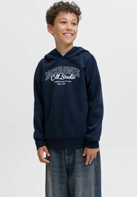 Lächelnder Junge mit lockigem Haar, der einen dunkelblauen Kapuzenpullover mit dem Aufdruck "JACK & JONES Coll. Studio" und lockeren blauen Jeans trägt, vor einem einfarbigen Hintergrund.
