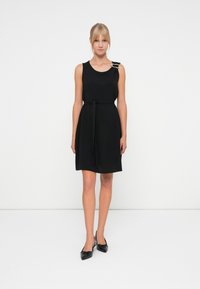 Robe noire sans manches avec une taille à nouer, présentant un décolleté arrondi et une boucle décorative sur une épaule. Texture de tissu lisse.