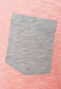 Rosa tygtextur med en grå fickdetalj. Fickan är rektangulär med en lätt melerad utseende och sydda kanter.