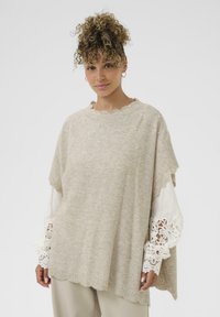 Poncho en tricot beige clair avec des bords festonnés, orné de petites perles, porté par-dessus un chemisier blanc à manches longues avec des poignets en dentelle.