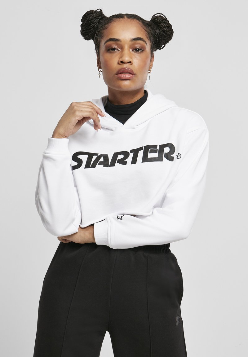 Felpa corta bianca con logo "STARTER" in nero, dotata di tasca frontale e cappuccio con cordino. Abbinata a pantaloni della tuta neri.