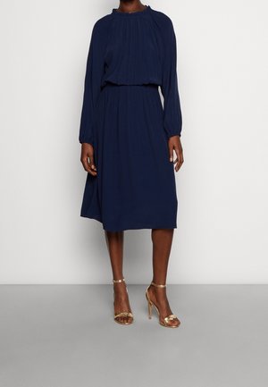 Kobieta w granatowej, plisowanej sukience midi z długim rękawem i złotych sandałach na wysokim obcasie, stojąca na jednolitym tle.