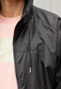 Person som bär en svart zipjacka med huva och dragsko över en rosa skjorta med delvis synlig ljusfärgad text.