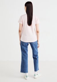 Femme avec de longs cheveux raides portant un t-shirt rose clair, un jean bleu, des chaussettes blanches et des baskets blanches avec des accents verts, se tenant devant un fond blanc.