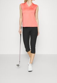 Korallfärgad ärmlös polo med krage, matchad med svarta capribyxor med sidslitsar. Bärs med vita golfsko och håller i en golfklubba.