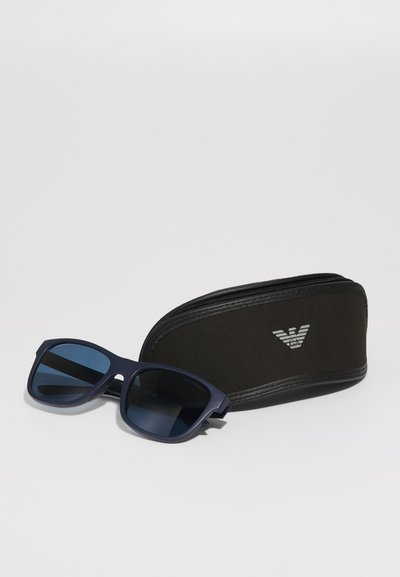 Ochelari de soare navy cu lentile întunecate, ramă pătrată și finisaj mat, alături de un toc negru cu fermoar care prezintă un logo.