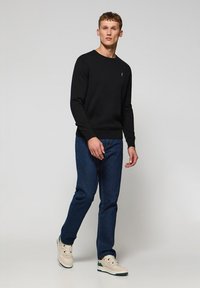Sudadera negra de cuello redondo, vaqueros de denim azul y zapatillas beige con acentos verdes. El modelo está de pie contra un fondo neutro.