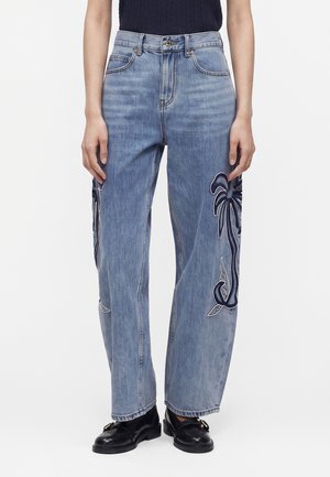 Kobieta nosząca niebieskie jeansy z wysokim stanem, z ciemnym haftem z palm na nogawkach, połączone z czarnymi mokasynami i ciemnym, bezrękawnym topem.