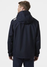 Marineblauwe waterdichte jack met capuchon, gestructureerde stof en witte logo-accenten op de schouders, ontworpen voor buitengebruik.
