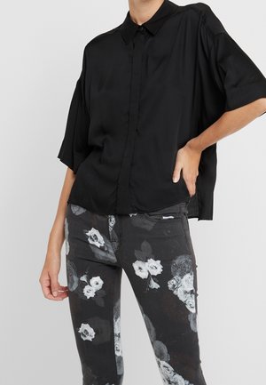 Person trägt ein lockeres schwarzes kurzärmeliges Hemd mit Knöpfen und schwarze Hose mit weißen und grauen Blumenmustern, steht mit einer Hand in der Hüfte.