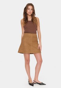 Jupe mini en daim marron avec une forme évasée et des détails cousus. Associée à un débardeur ajusté marron et des escarpins noirs à bout pointu.