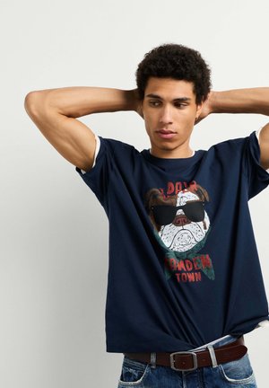 Joven con pelo rizado que lleva una camiseta azul marino con la cara de un perro con gafas de sol y texto rojo, de pie con las manos detrás de la cabeza.