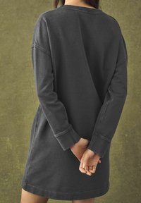 Robe sweat-shirt noire, surdimensionnée, avec manches longues et encolure ronde. Présente une texture subtile et des poignets côtelés, portée de manière décontractée.