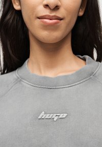 Κοντινό πλάνο ενός ατόμου που φοράει γκρι φούτερ με λευκή κεντημένη λέξη «hergo» στο στήθος.