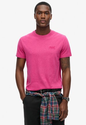 Lyserød t-shirt med korte ærmer og rund halsudskæring samt broderet logo. Båret med et sort bælte og en ternet skjorte knyttet om taljen.