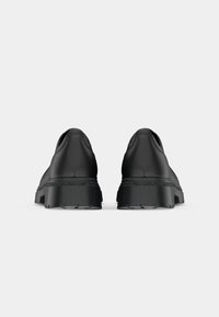 Zapatos negros tipo slip-on con un acabado de cuero suave, suela de goma gruesa y detalles texturizados, presentan una puntera redondeada y un diseño minimalista.