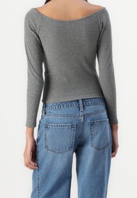 Persona che indossa un top grigio attillato a spalle scoperte e jeans blu in denim larghi, vista da dietro su uno sfondo semplice.