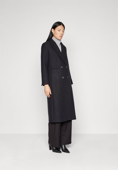 Cappotto nero doppiopetto con rever oversize, tasche laterali e una silhouette lunga, abbinato a pantaloni larghi scuri e tacchi a punta neri.
