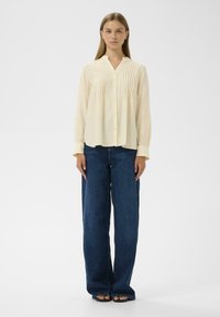Lichtgele button-up blouse met verticale plooien, v-hals en lange mouwen; gecombineerd met hoogwaist donkerblauwe wijde jeans.