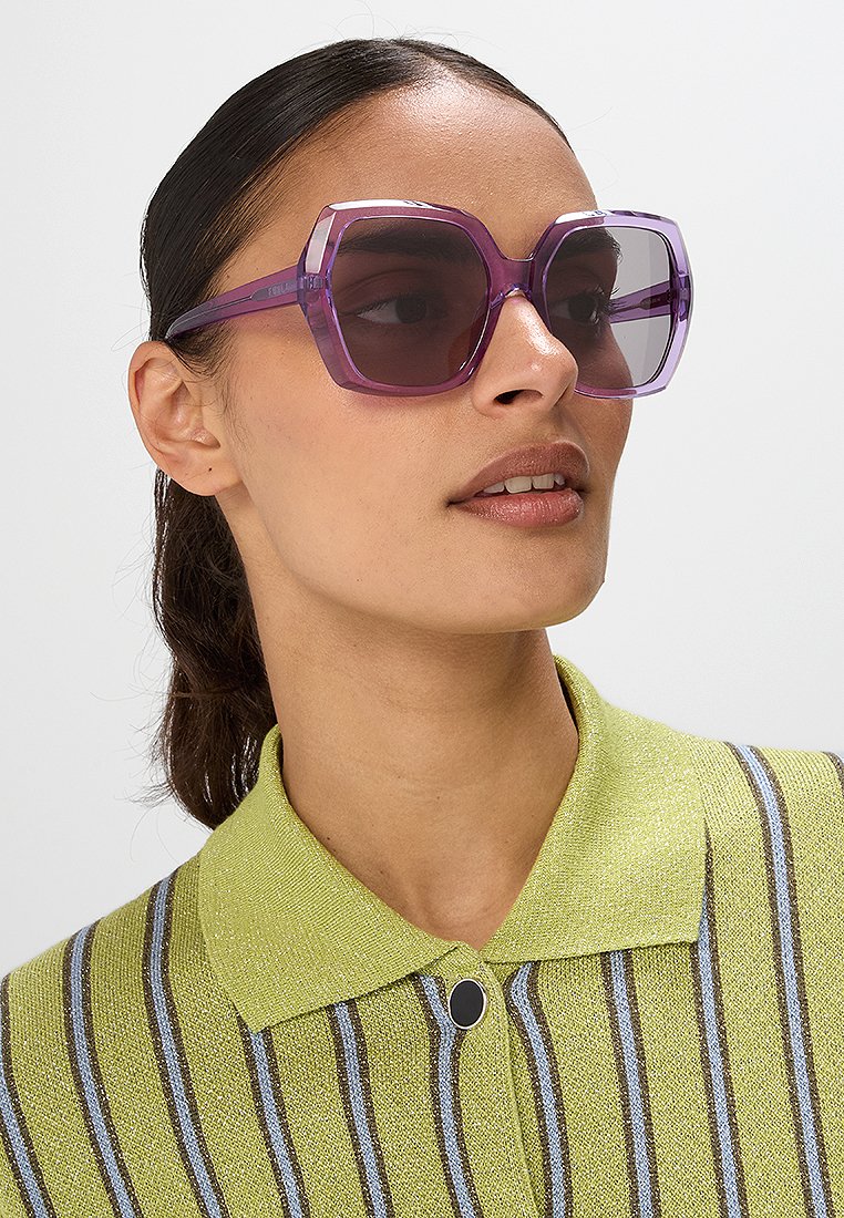 Occhiali da sole oversize viola a forma esagonale, con montatura traslucida e lenti scure. Il modello indossa un maglione a righe verdi con colletto.