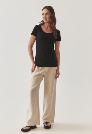 T-shirt noir à manches courtes avec un col dégagé associé à un pantalon large en lin beige clair et des sandales glissantes noires.