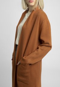 Kobieta w beżowej bluzce i długim brązowym kardiganie z kieszeniami, pozująca z jedną ręką w kieszeni, na jednolitym jasnym tle.