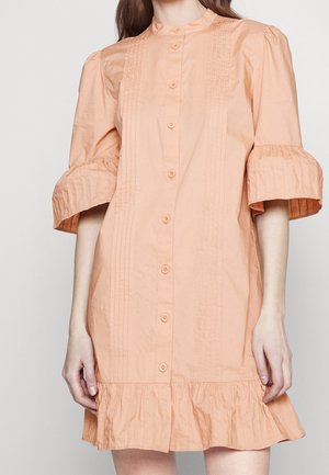 Kobieta w brzoskwiniowej sukience zapinanej na guziki, z bufkami sięgającymi do łokcia oraz plisowanymi detalami z przodu i na dole sukienki.