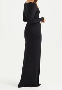 Kvinna i en lång, åtsittande svart klänning med långa ärmar, som står med ryggen och sidan synlig mot en plain vit bakgrund.