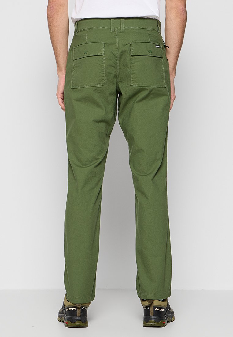 Grüne Baumwollhose mit geraden Beinen, ausgestattet mit zwei Gesäßtaschen und einem Knopfverschluss. Trägt dazu schwarze und grüne Sportschuhe.