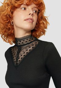 Schwarzes, geripptes Oberteil mit hohem Spitzenkragen, der geometrische Muster und gewellte Kanten aufweist und einen strukturierten Kontrast am Ausschnitt betont.