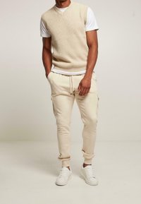 Beżowa kamizelka z prążkowanego dzianiny na białej koszulce, w połączeniu z beżowymi spodniami dresowymi z kieszeniami cargo i białymi sneakersami. Prosty fason.