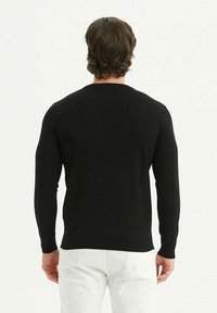Pull en tricot noir à manches longues, col rond et poignets côtelés. Le tissu semble lisse et extensible. Porté avec un pantalon blanc.