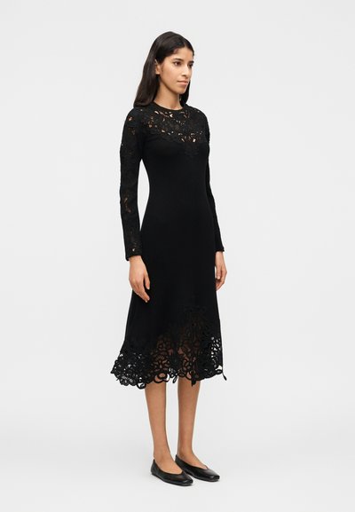Γυναίκα που φοράει μαύρο midi φόρεμα με μακριά μανίκια και δαντελένιες λεπτομέρειες στο στήθος, στα μανίκια και στο τελείωμα, συνδυασμένο με μαύρες μπαλαρίνες, στέκεται πλαγίως.