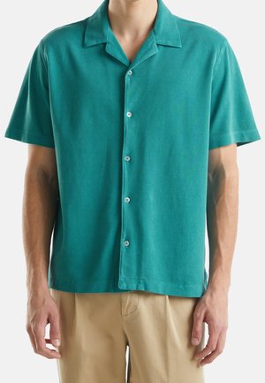 Mies, jolla on päällään petroolinvihreä lyhythihainen nappipaita, jossa on camp-kaulus, ja beiget housut, seisoo käsivarret rentoina sivuilla.