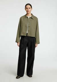 Chaqueta corta verde oliva con cuello de solapa, cierre frontal de botones y dos bolsillos laterales; combinada con pantalones de pierna ancha negros.