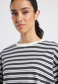 Jonge vrouw met donker haar dat in een staart is geknoopt, draagt kleine gouden oorbellen en een wit shirt met horizontale zwarte strepen, met een neutrale uitdrukking, op een witte achtergrond.