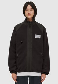 Zwarte fleece jas met een hoge kraag, contrasterende schouderpanelen, ritssluiting aan de voorkant en zijzakken. Bevat een logo patch op de borst.