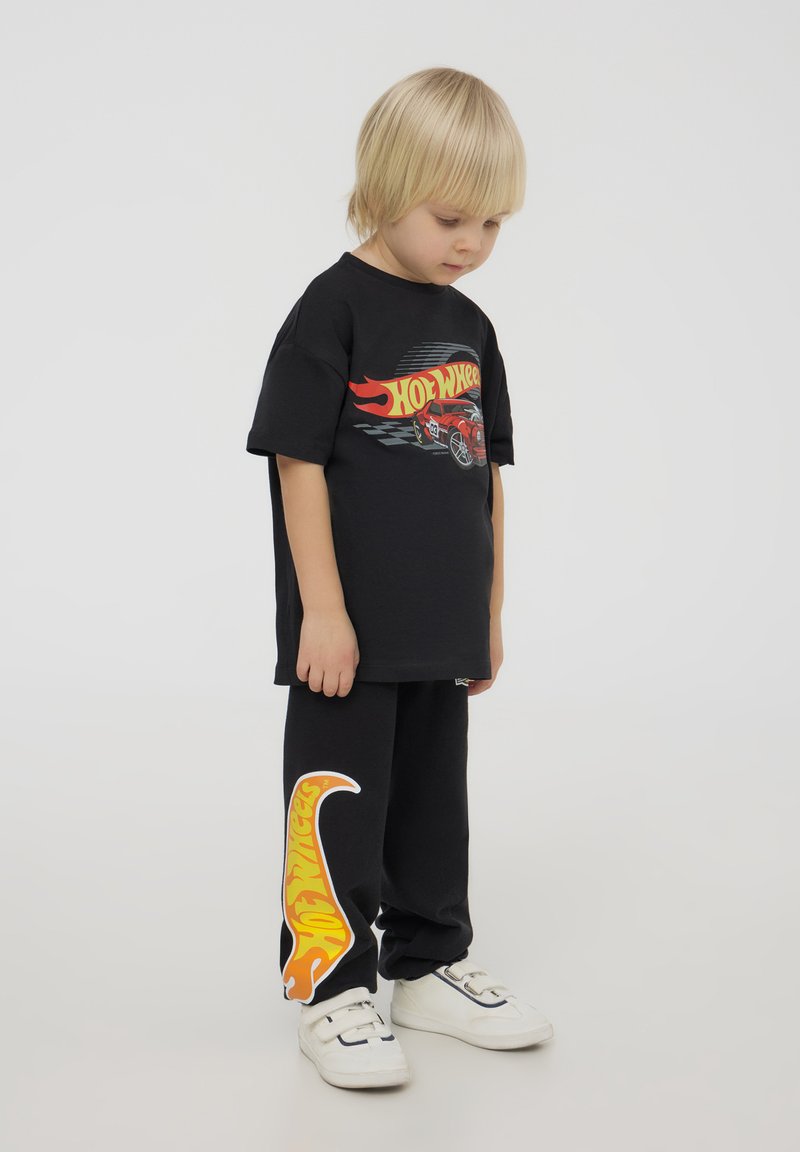 Maglietta in cotone nera con grafica "Hot Wheels" rossa e gialla e dettagli a scacchi neri, abbinata a pantaloni neri con logo a fiamma gialla.