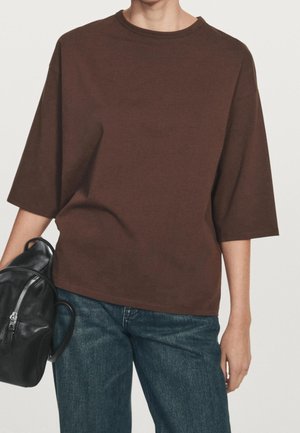 Personne portant une chemise marron ample à manches courtes et un jean bleu, tenant un sac à dos en cuir noir par une poignée.