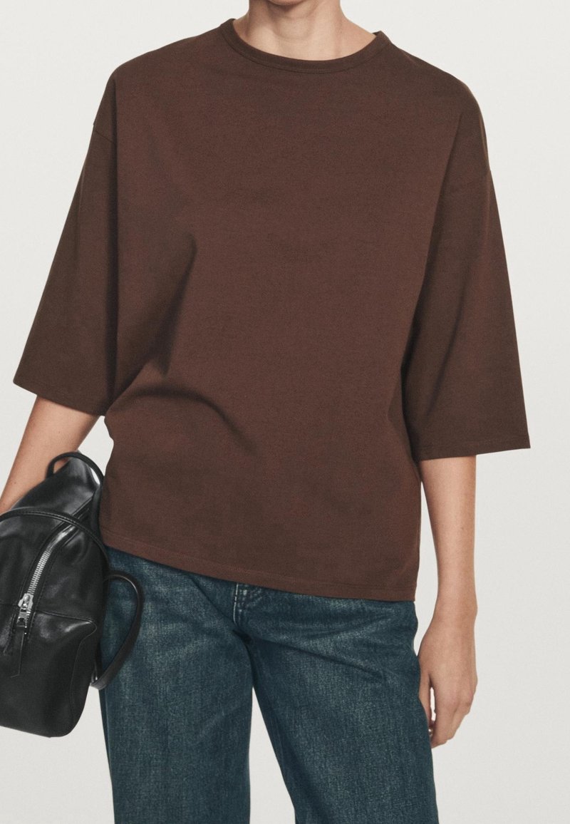 Personne portant une chemise marron ample à manches courtes et un jean bleu, tenant un sac à dos en cuir noir par une poignée.