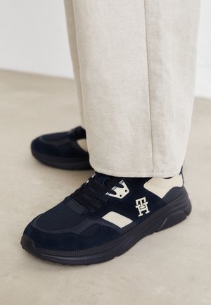 Granatowo-beżowy sneaker z logo po boku, noszony z jasnobeżowymi spodniami, stojący na gładkiej jasnoszarej podłodze.
