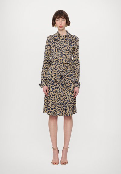 Long-sleeved, button-up dress in a beige and navy abstract pattern. A-line silhouette with a cinched waist and wrist ruffles. Sandals visible.