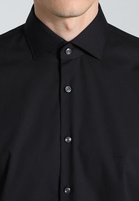 Svart skjorte med spiss krage, seks knapper og subtil logo-brodering. Glatt, rynkefritt stoff av middels vekt.