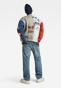 Färggad lapptäckshoodie med blå, röd och beige sektioner och grafiska tryck; blå jeans och vita sneakers fulländar looken.