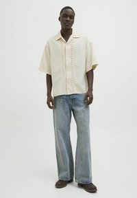 Camisa de botones de manga corta, de color claro, con perforaciones decorativas, combinada con vaqueros de pierna ancha azules y zapatos marrón oscuro.