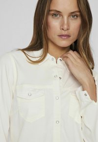 Camicia button-up a maniche lunghe di colore crema, con due tasche sul petto e chiusure a pressione. Tessuto liscio e design del colletto classico.