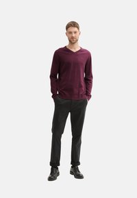 Suéter burdeos con cuello en V, hecho de un suave tejido de punto, combinado con pantalones negros y zapatos de cordones negros, de pie sobre un fondo neutro.